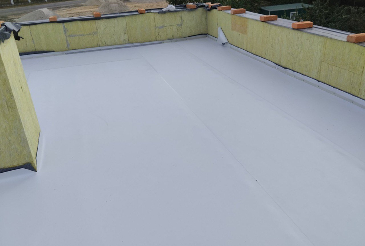 Flat rooftop with a smooth, white waterproof membrane installed, surrounded by yellow insulation panels and orange weights along the edges.