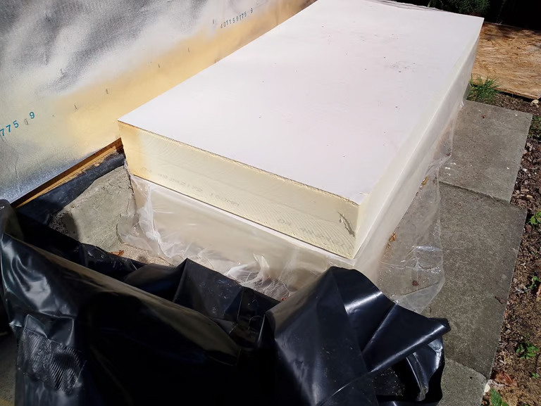 A large rectangular sheet of rigid foam insulation rests on a plastic-covered concrete surface outdoors, next to a wall with a partially installed vapor barrier.