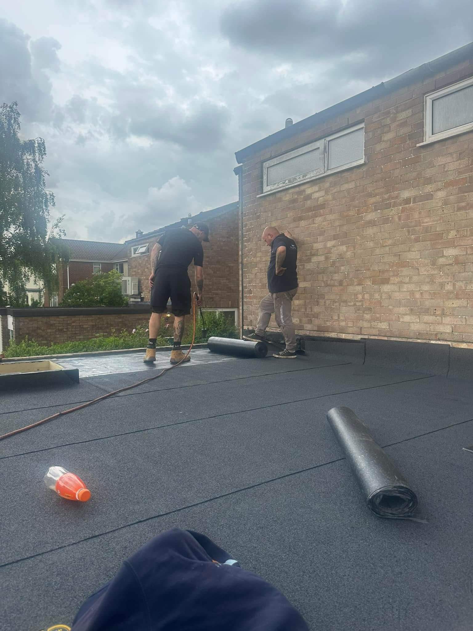 Two people are installing roofing material on a flat roof next to a brick building, with tools and rolls of material visible on the surface.