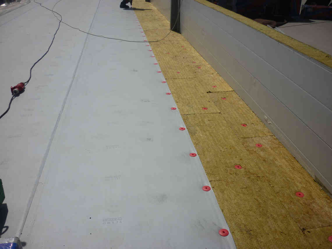 Flat roof under construction showing insulation boards on the right and membrane fastened with red washers on the left, with a tool and cable visible.