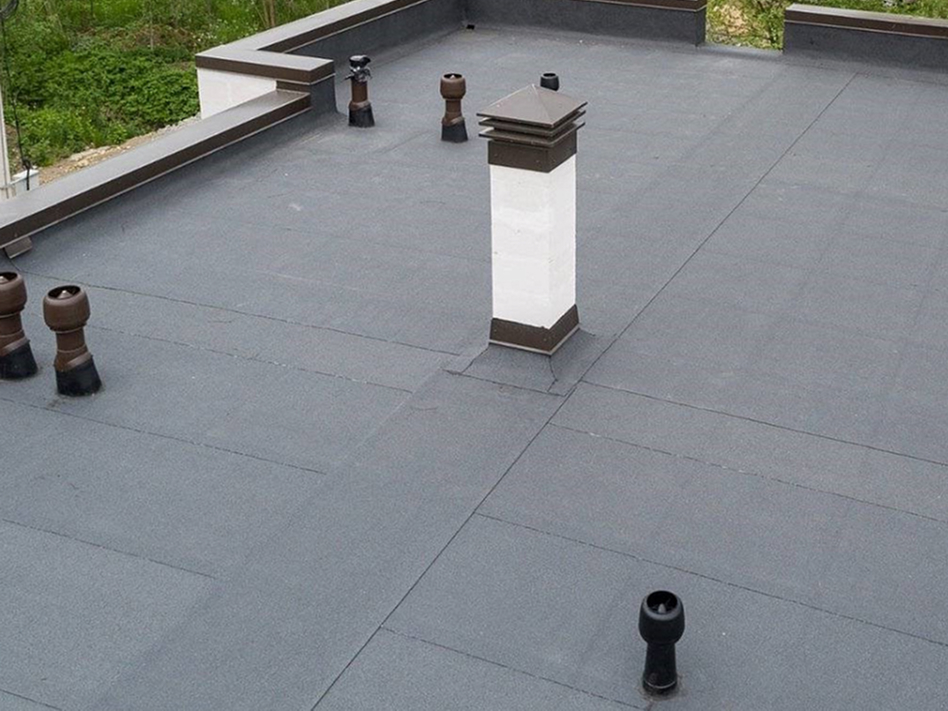 A flat rooftop with a gray waterproof membrane, several black vent pipes, and a white rectangular chimney with a dark cap.