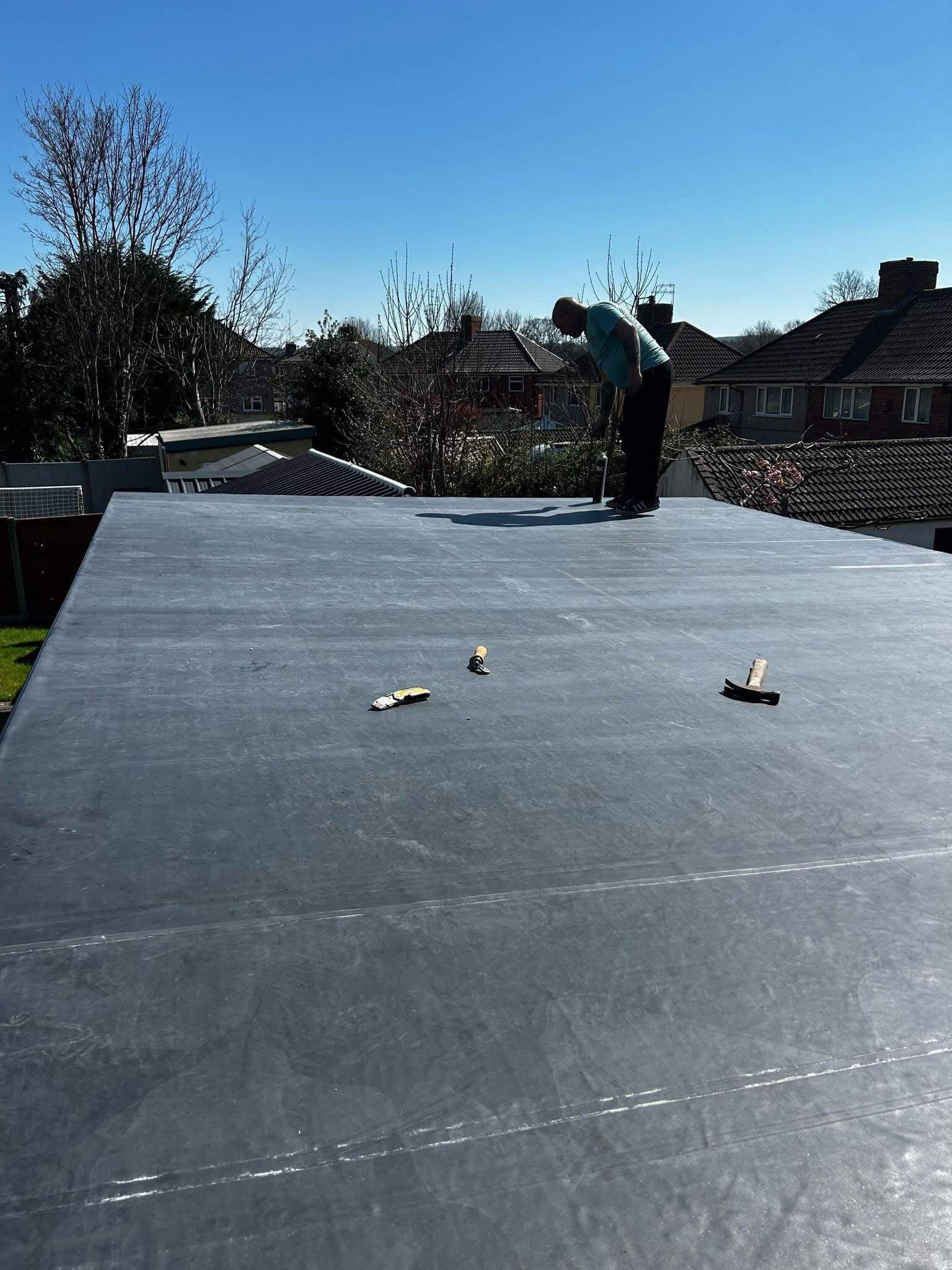 A person stands on a flat rooftop covered with black waterproof material, with tools scattered on the surface. Houses and trees are visible in the background under a clear sky.